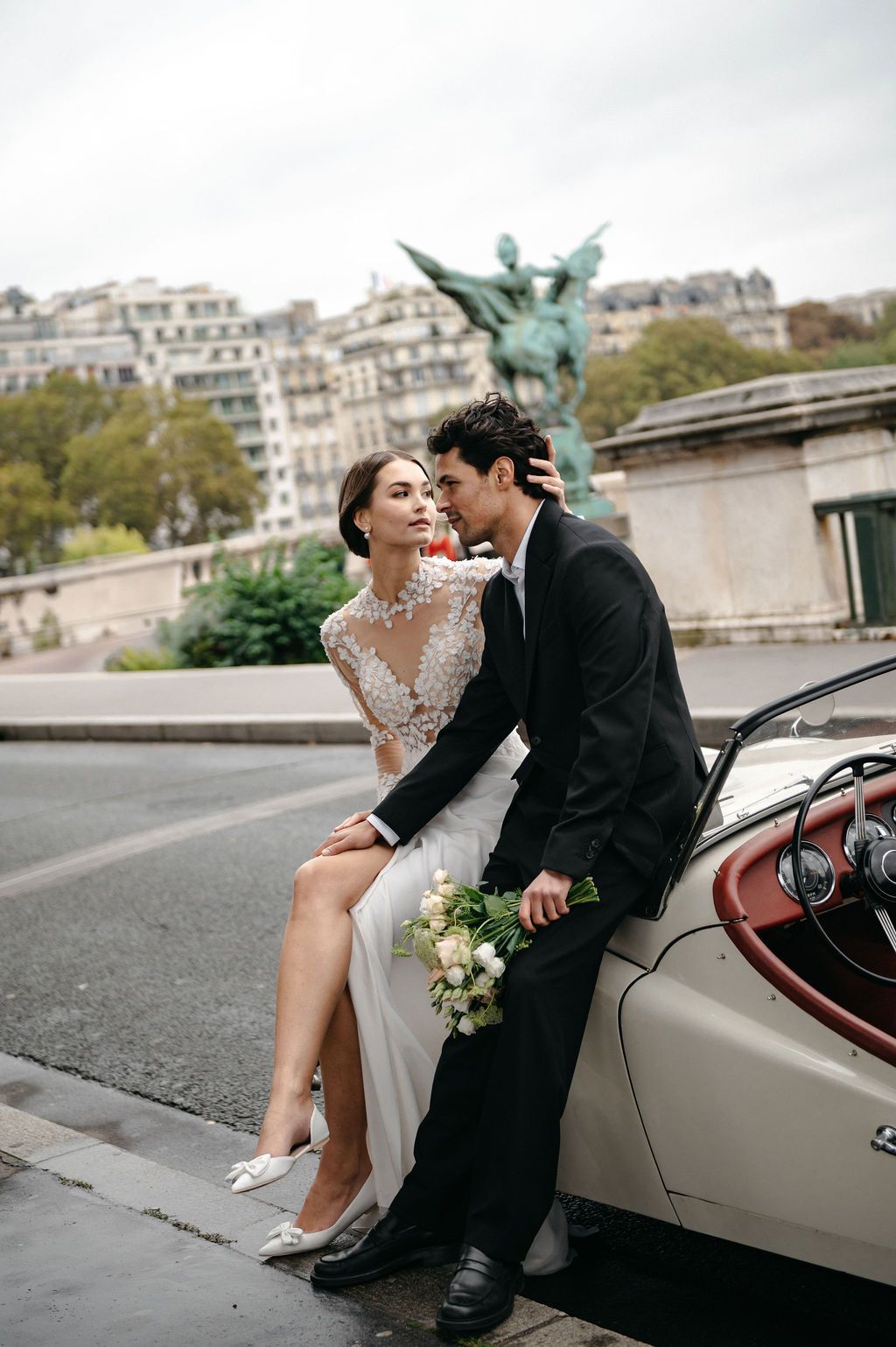 Bride in lace gown and groom in black suit pose by vintage car in Paris with statue backdrop.