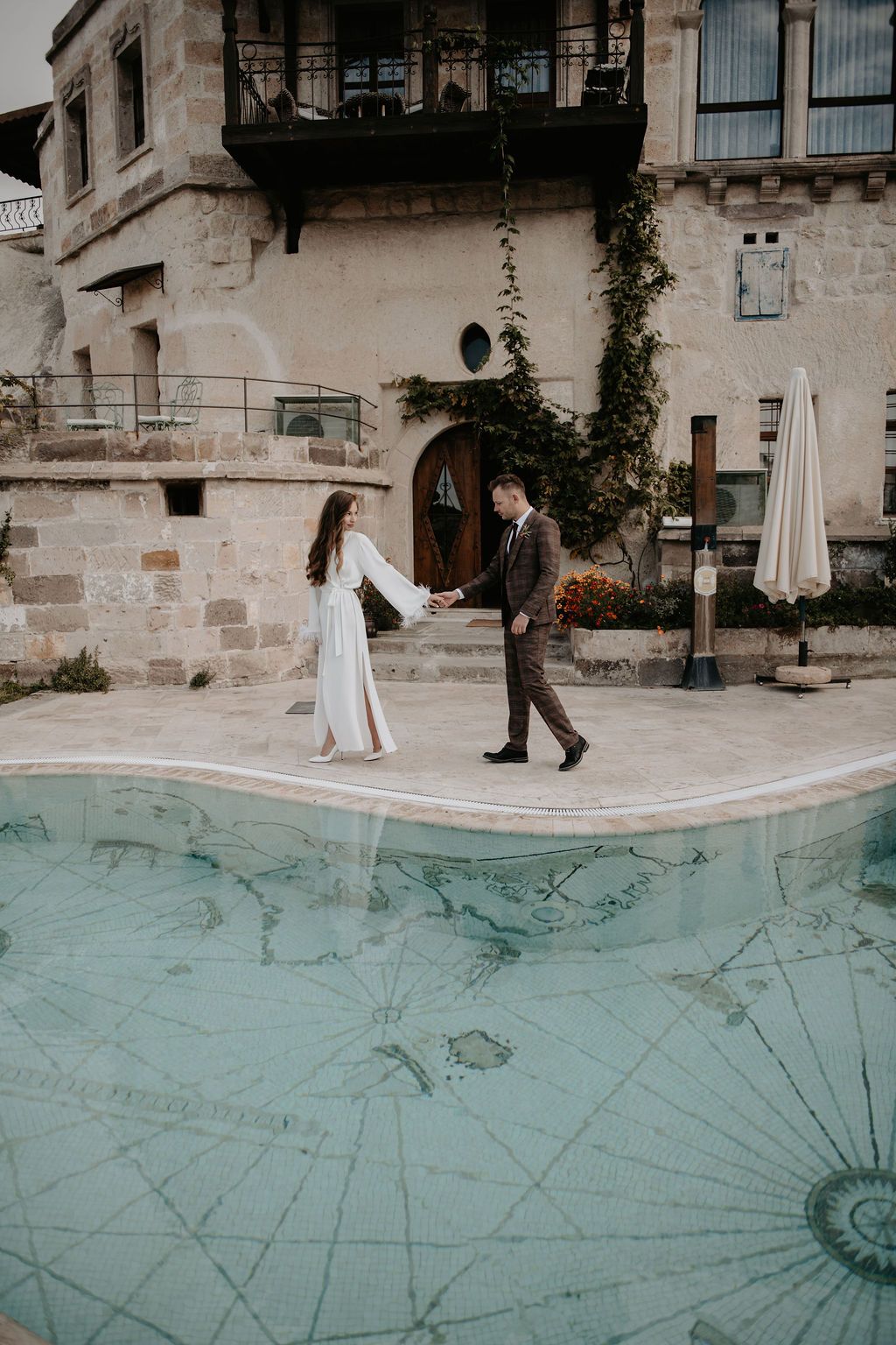 Couple holding hands walking beside a map-tiled pool at a stone villa in Cappadocia, Turkey.