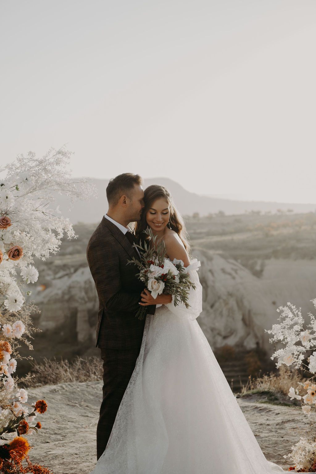 Bride and groom embracing at outdoor wedding ceremony with floral arch and scenic landscape.
