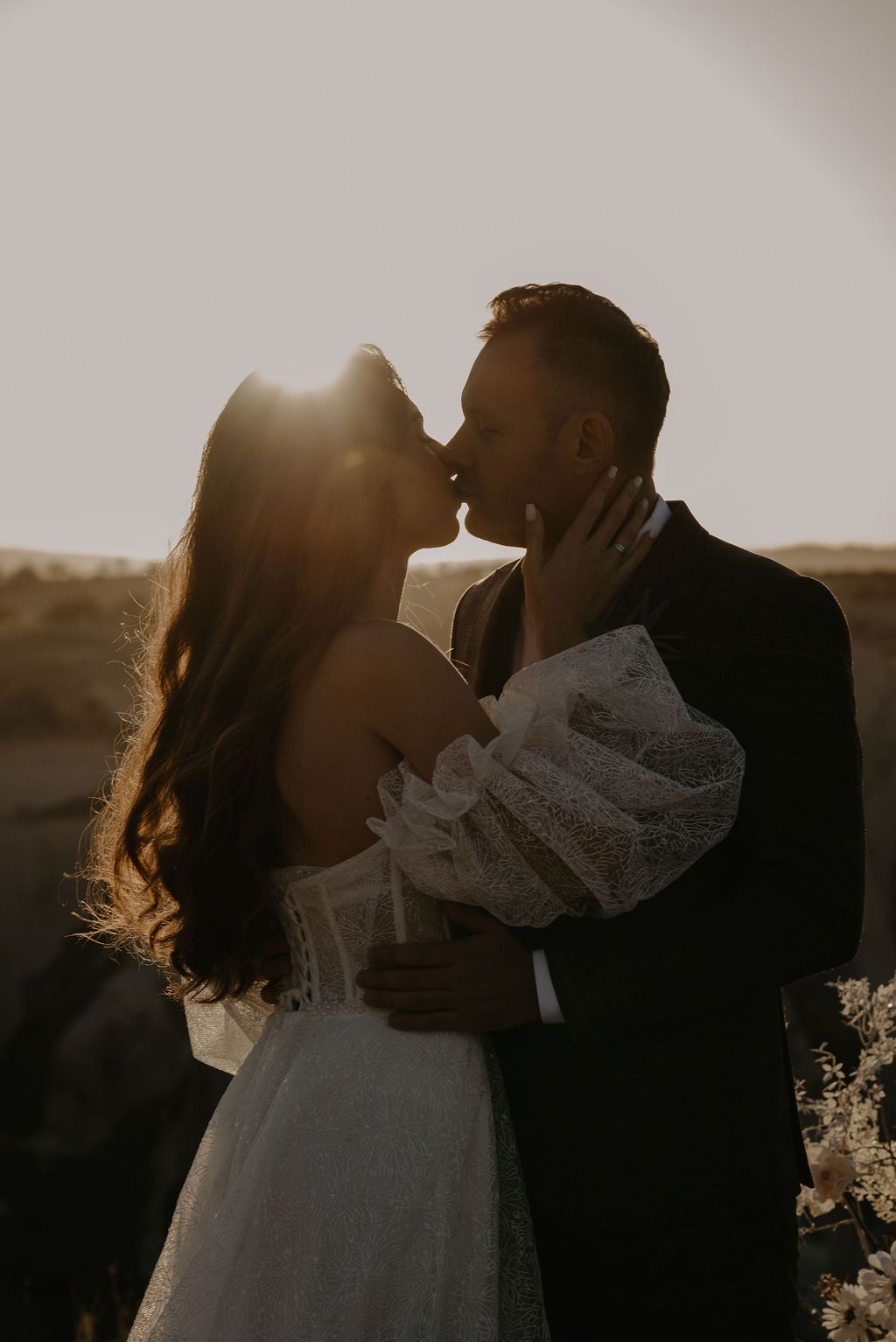 Couple shares a romantic sunset kiss, bride in lace off-shoulder gown holding a bouquet.