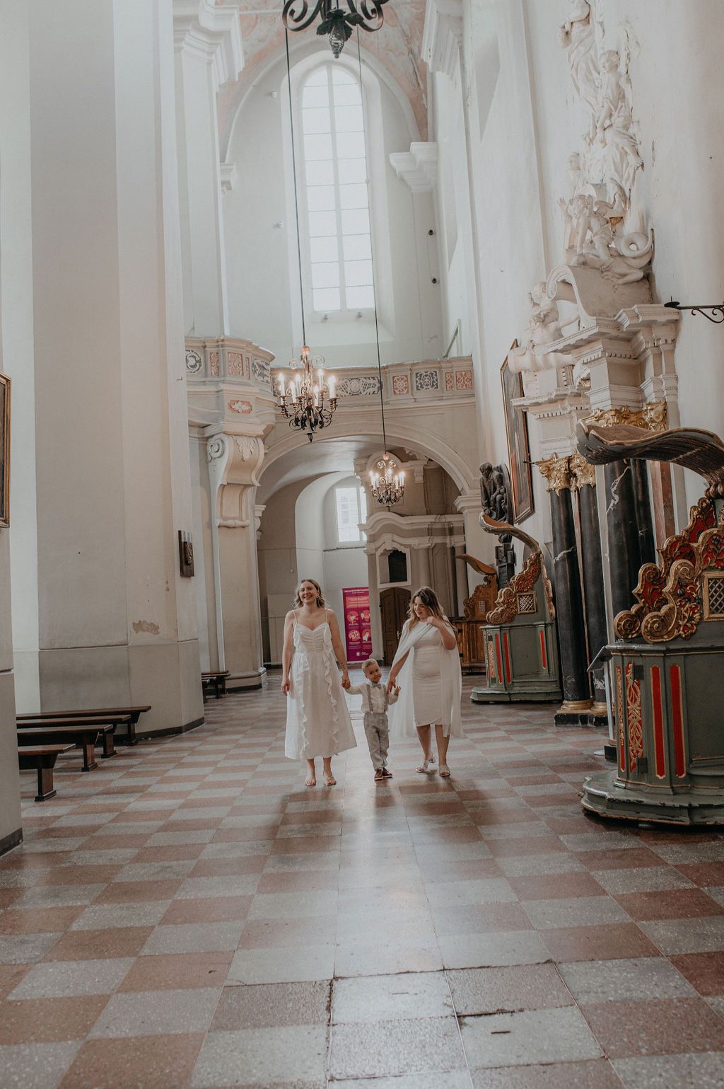 Two women in white dresses walk with a child through an ornate European church interior with chandeliers.