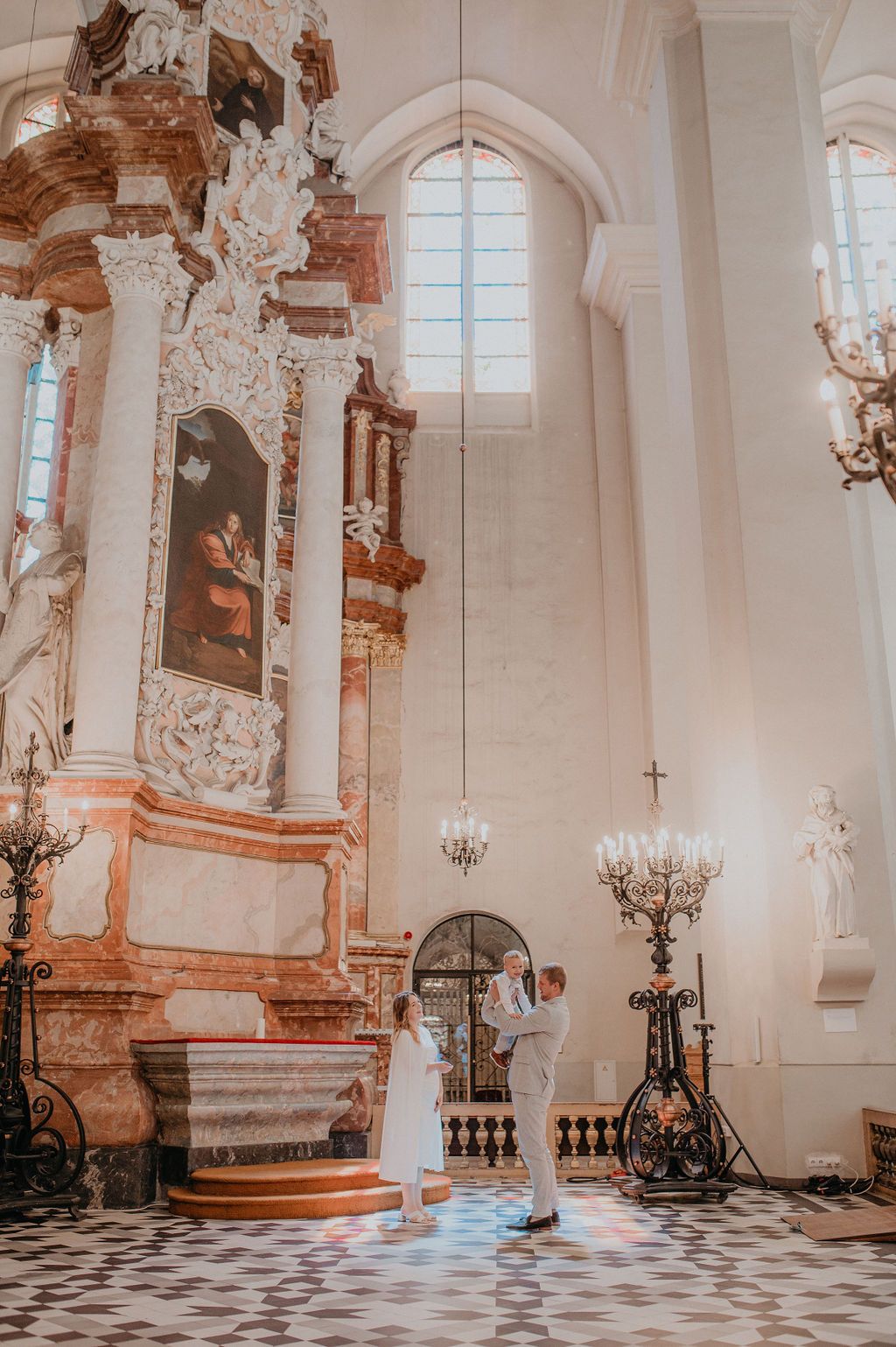 Couple sharing intimate moment at ornate baroque church altar with chandelier and classical paintings.