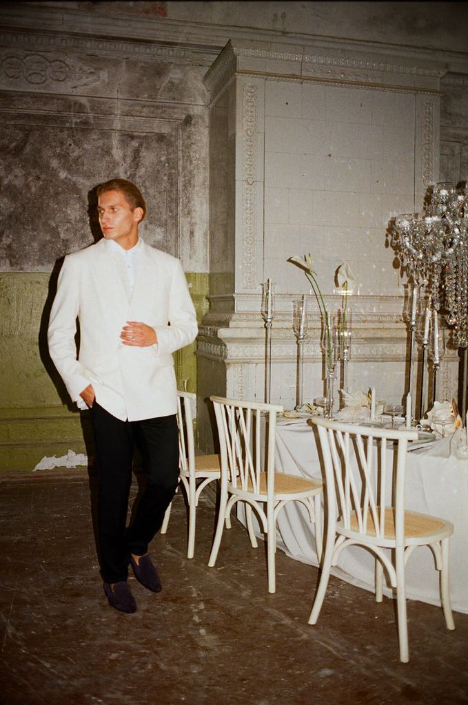 Man in white jacket and black pants stands beside an elegant table with candelabras in a vintage hall.