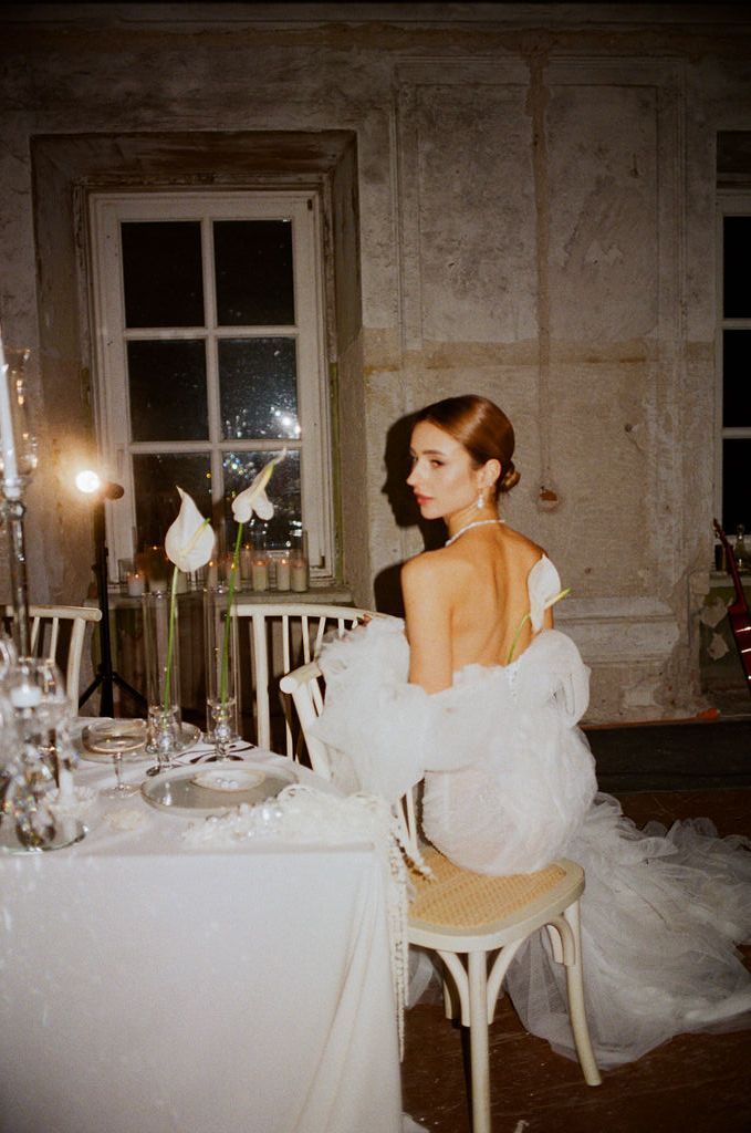 Elegant bride in white gown seated at candlelit reception table in rustic venue, gazing away romantically.