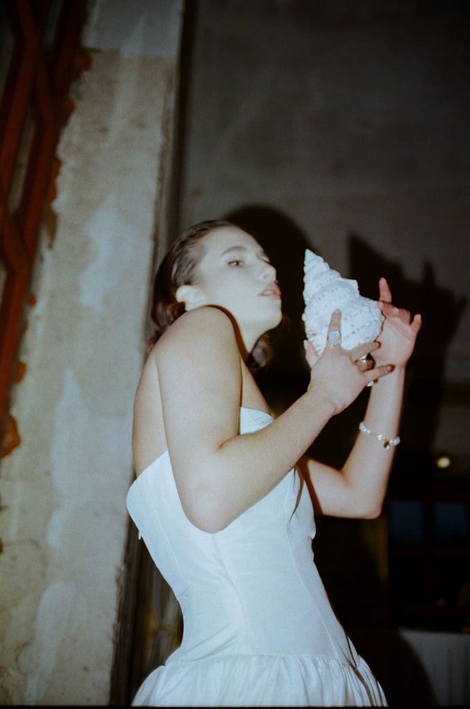 A young woman in a white strapless dress holds a large conch shell to her lips in a dimly lit room.