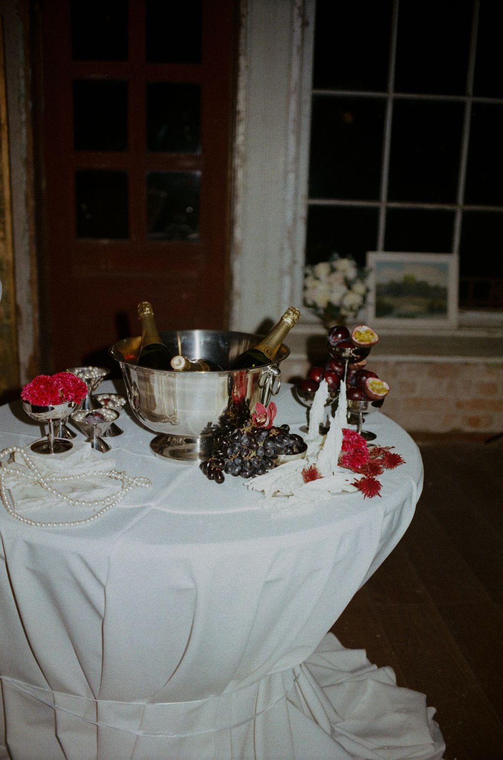 Elegant wedding cocktail table with white linen, champagne bucket, grapes, pearl necklace, and floral decorations at night.