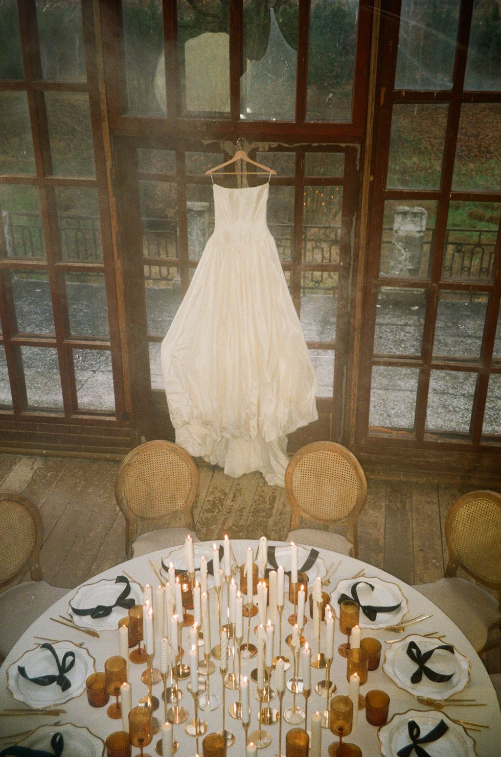 White wedding dress hanging above an elegantly set round table with candles and black ribbon napkins in a rustic venue.