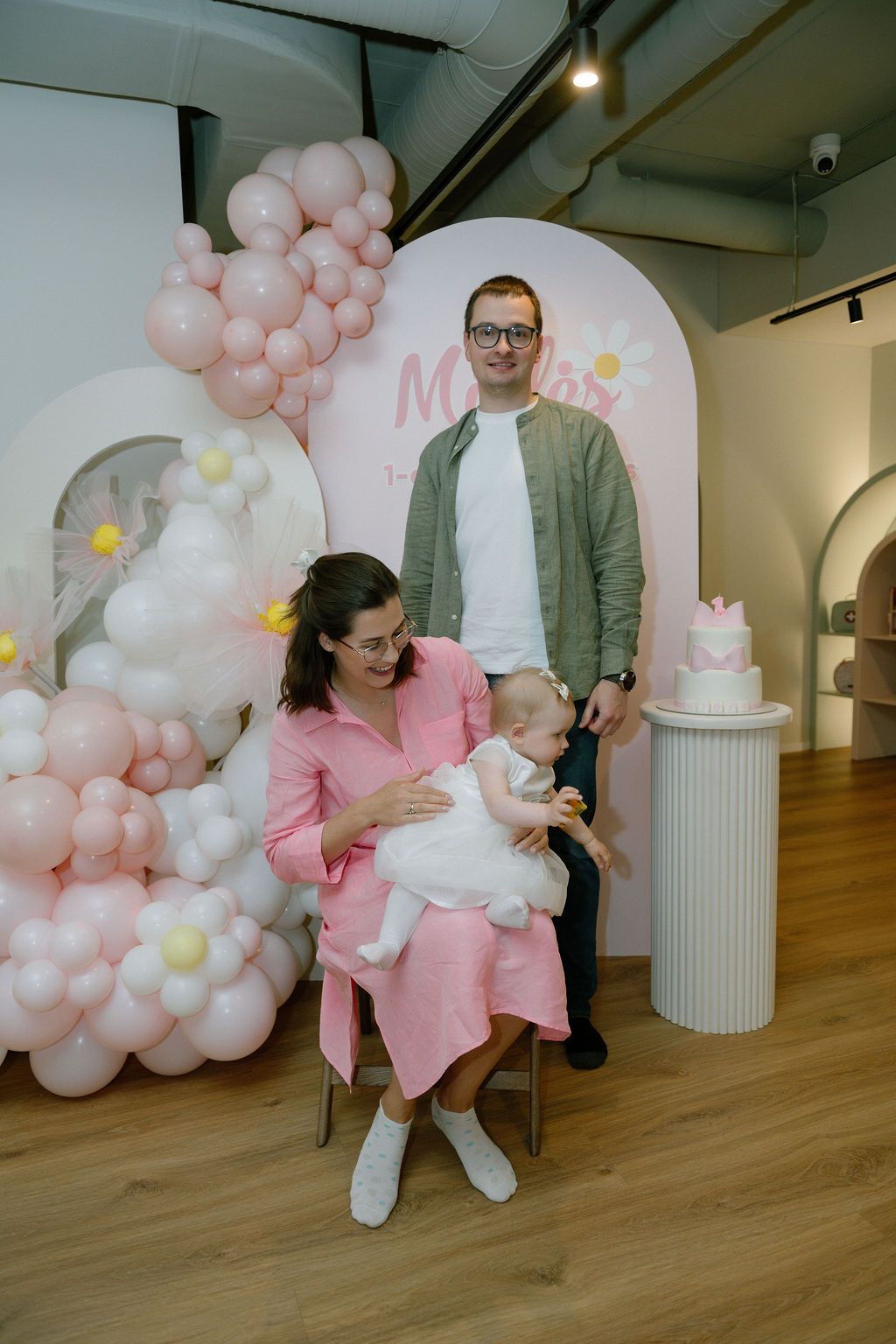 Family posing at a pink balloon-decorated birthday party backdrop with a baby in white dress and pink cake nearby.
