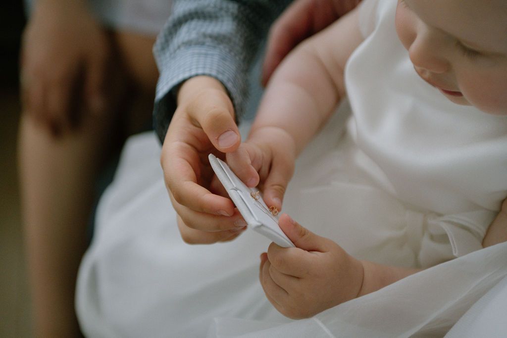 Adult hands helping a baby in white christening gown hold a small white card during a baptism ceremony.