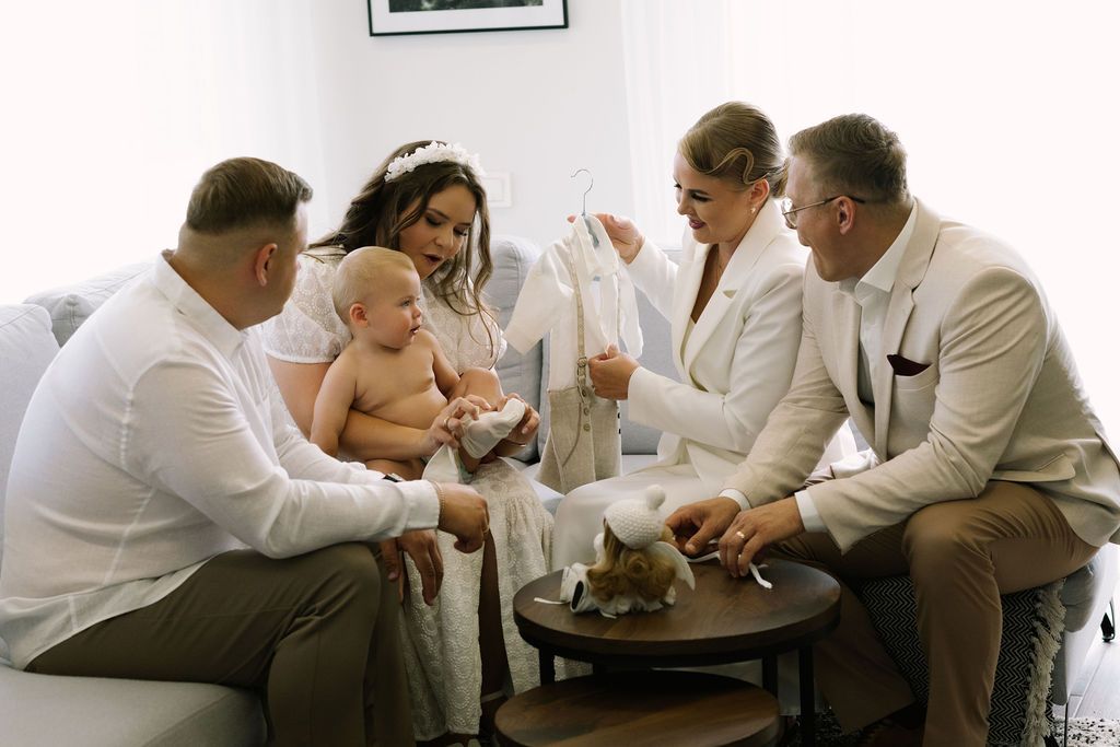 Family group in white attire gathered around a coffee table, admiring baby clothing at a christening.