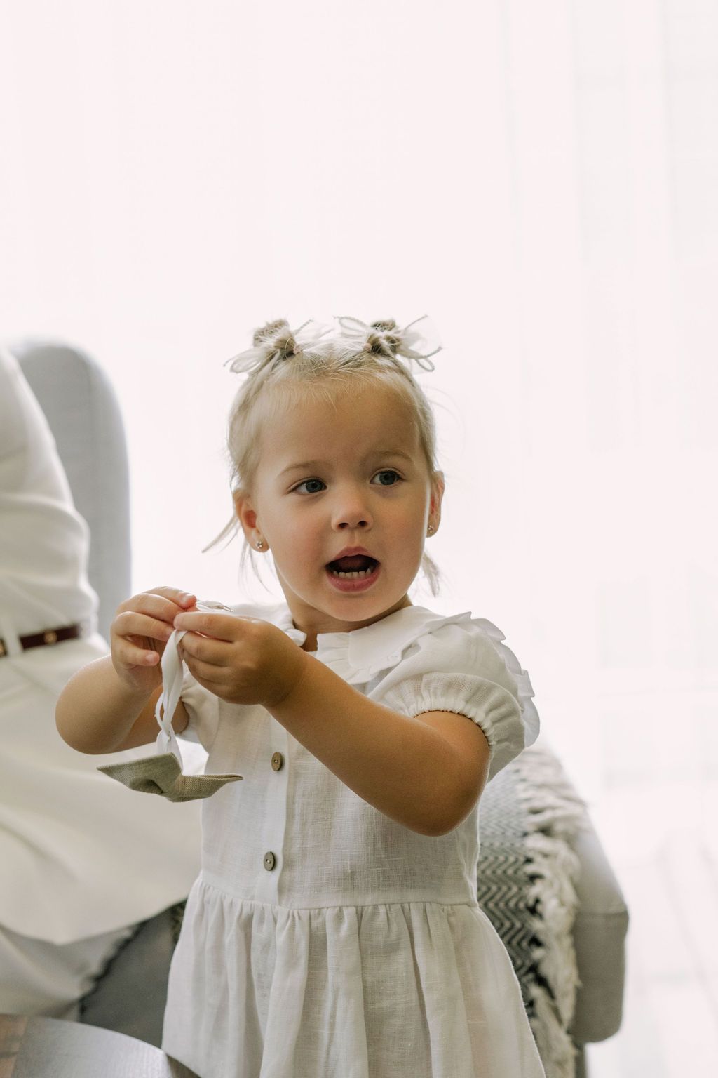 Adorable toddler girl in white linen dress with pigtails reaching out her hands indoors.