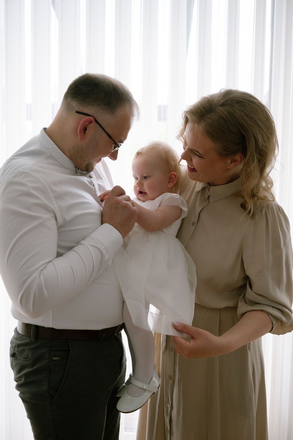 A happy family with a man in glasses, blonde woman in beige dress, and baby in white outfit standing by a bright window.
