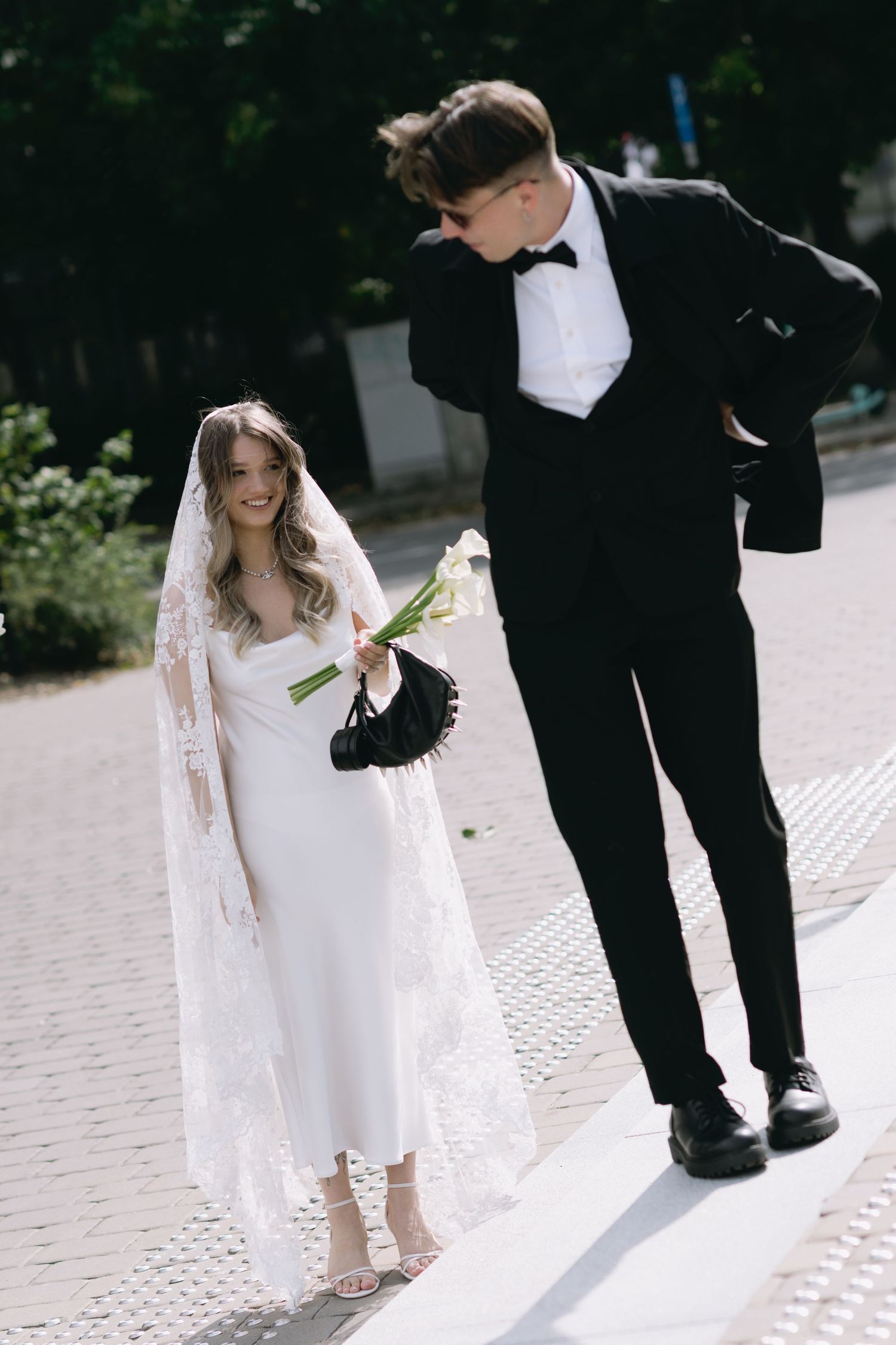 Bride in a white slip dress and lace veil holding calla lilies smiles as the groom in a black tuxedo and bow tie leans toward her.