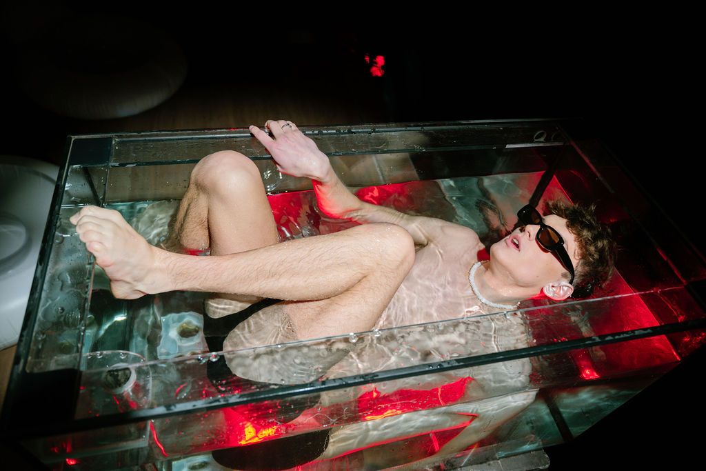 Person in sunglasses and a light outfit reclines inside a glass tank lit by red lights in a dark, moody room.