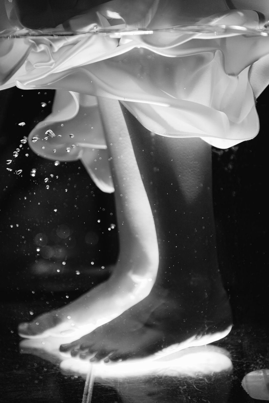 Black and white photo of bare feet standing underwater beneath a flowing white dress, with air bubbles rising in the dark water.
