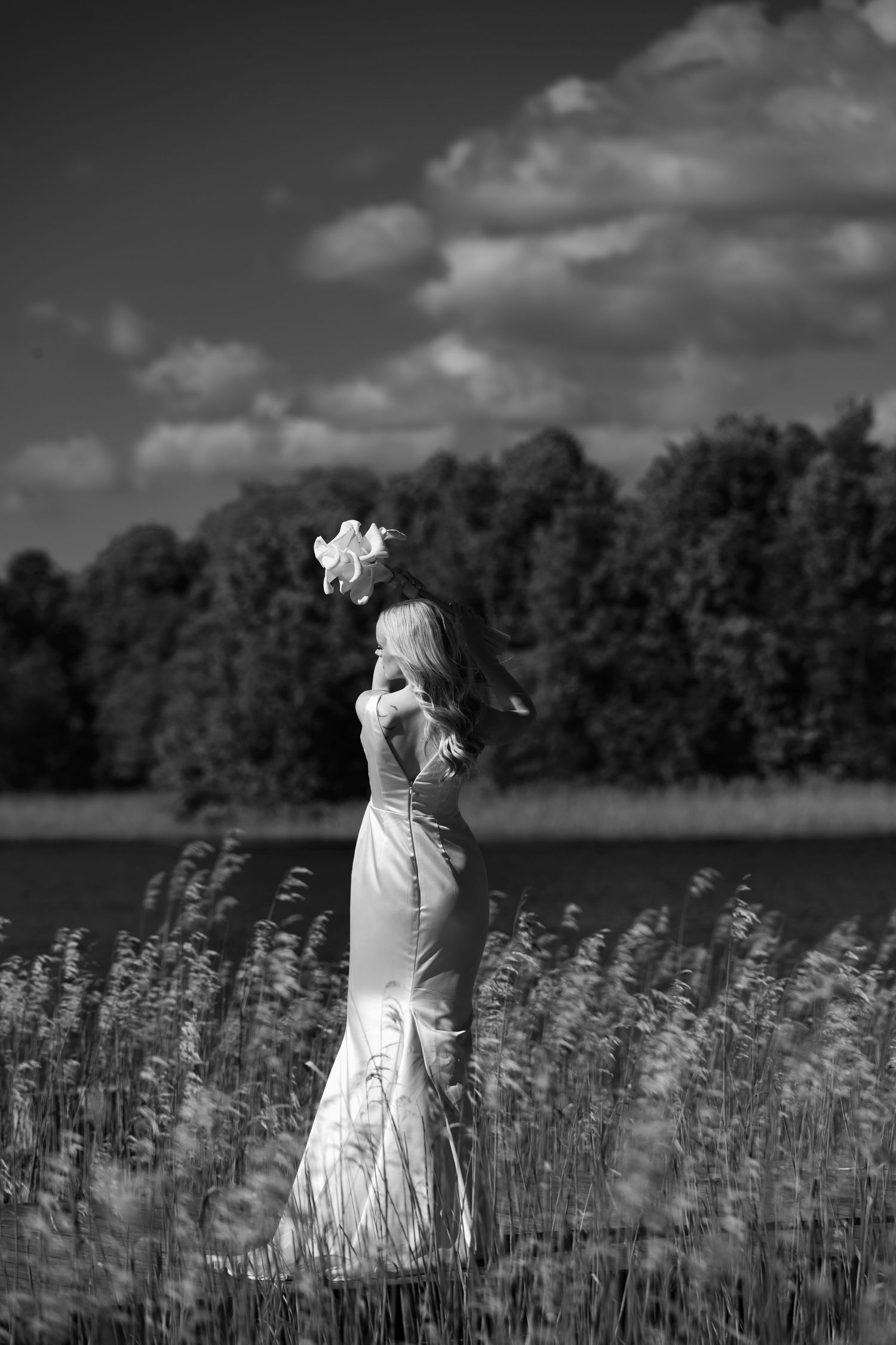Bride in a white gown and floral hair piece stands in tall reeds by a lake, in a black and white wedding portrait.