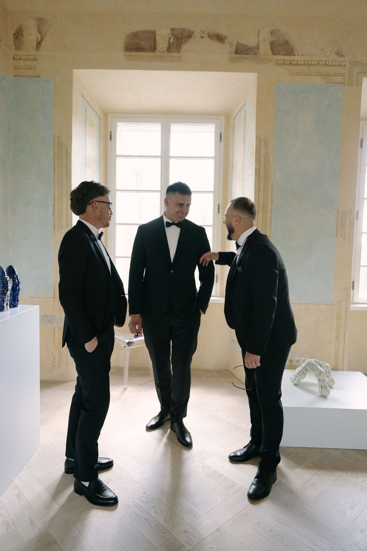 Three groomsmen in black suits chat and adjust their attire in a bright room with tall windows.