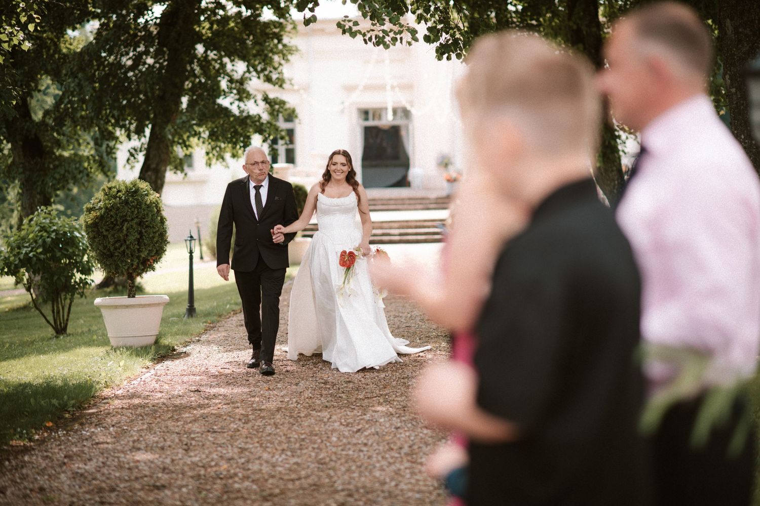 Bride in white gown walks with escort along tree-lined garden path toward wedding guests at elegant outdoor venue.
