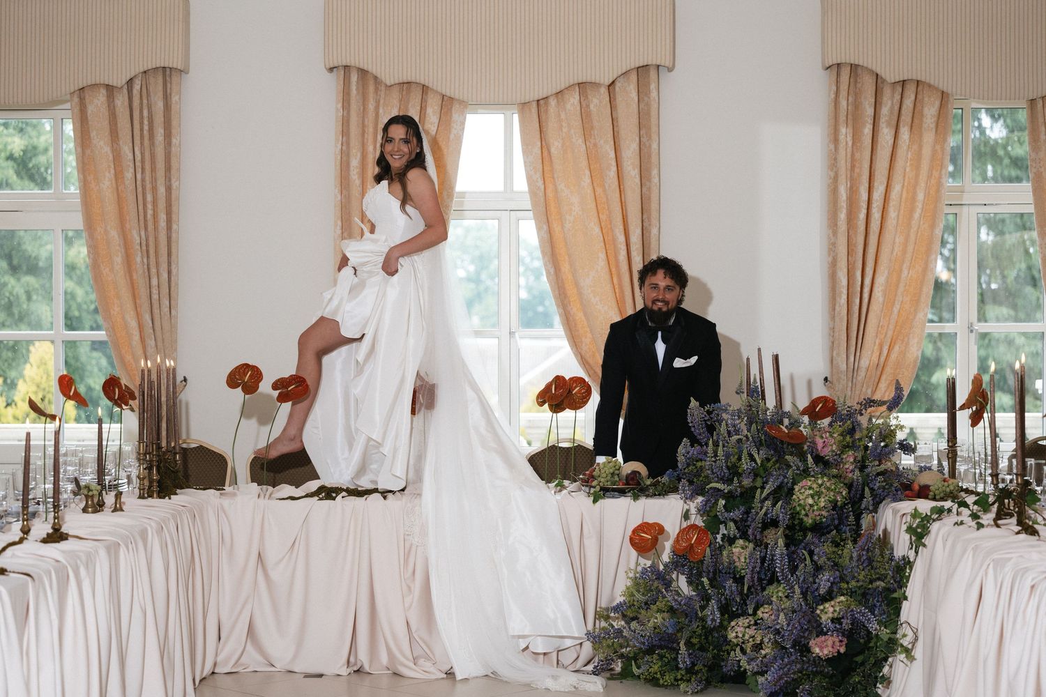 Elegant bride and groom posing at floral-adorned wedding reception table in a bright, curtained ballroom.