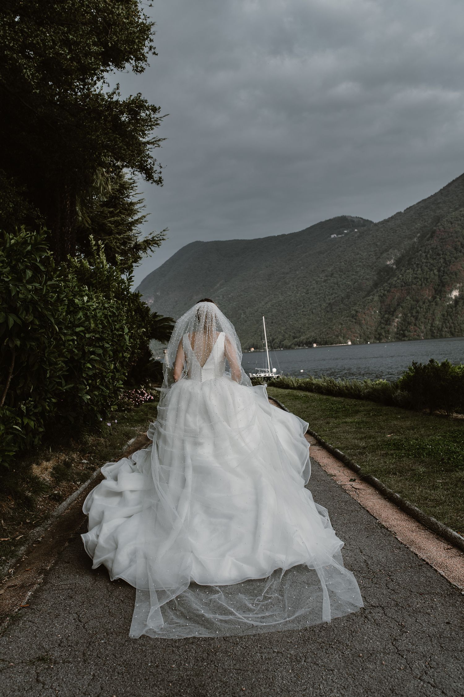 A bride in a flowing white wedding dress sits on a stone path overlooking misty mountains and water on an overcast day.