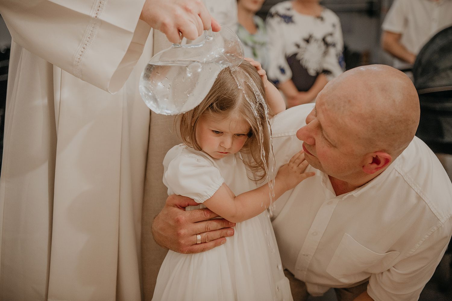 A tender moment during a baptism ceremony as water is poured over a child in a white dress.