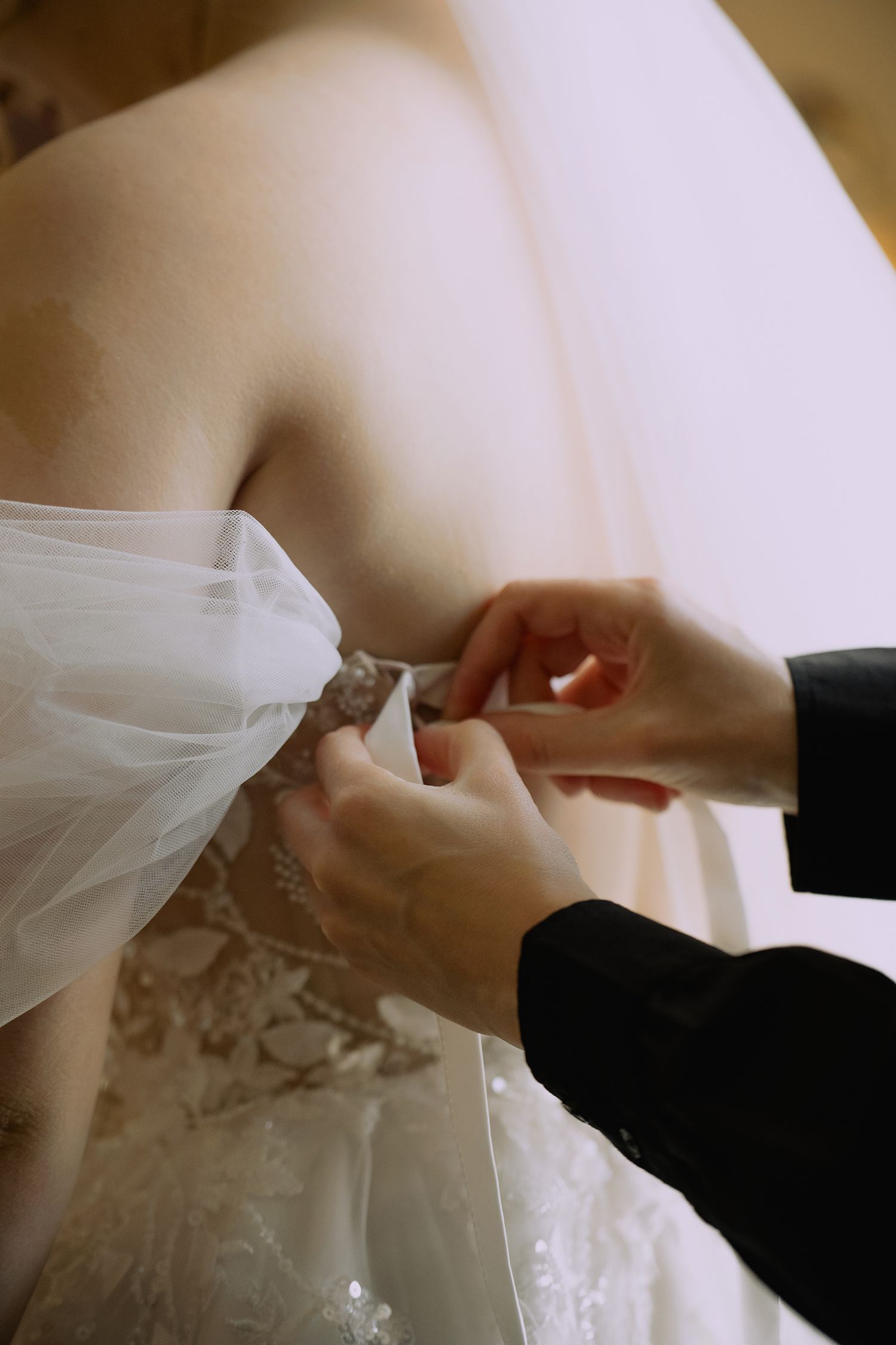Hands adjusting and fastening the delicate lace details on a white wedding dress.
