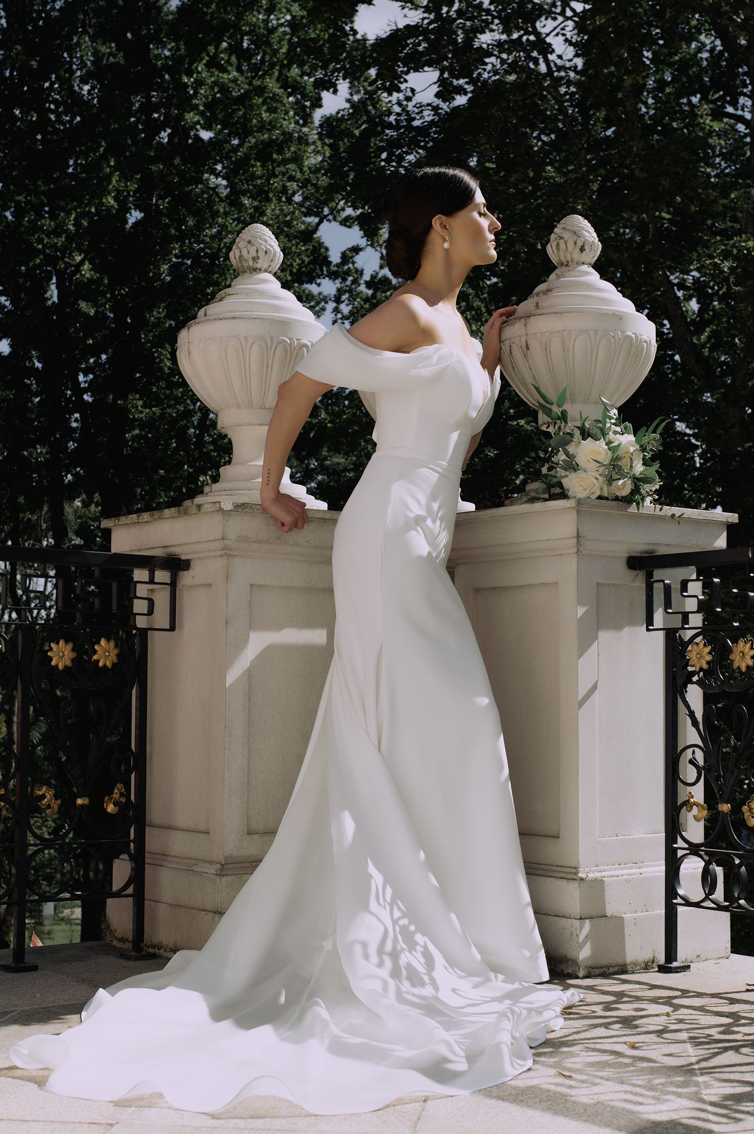 A person in a flowing white wedding dress poses elegantly against ornate stone pillars in an outdoor setting.