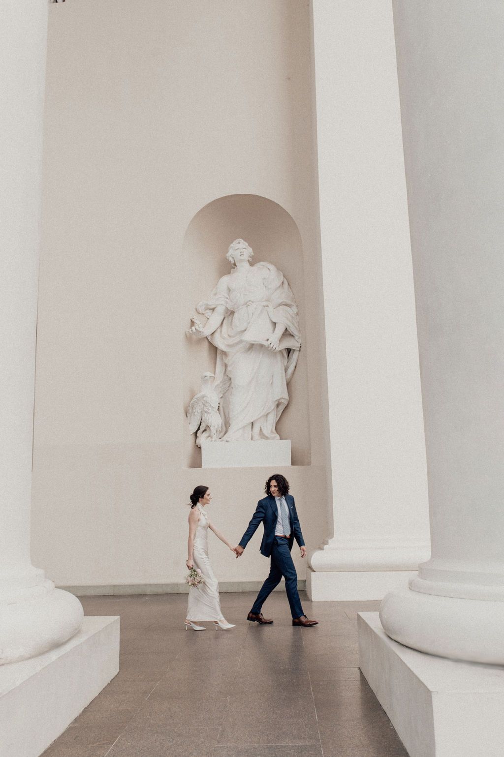 Bride and groom walk hand in hand past a classical white statue in an elegant columned hall.