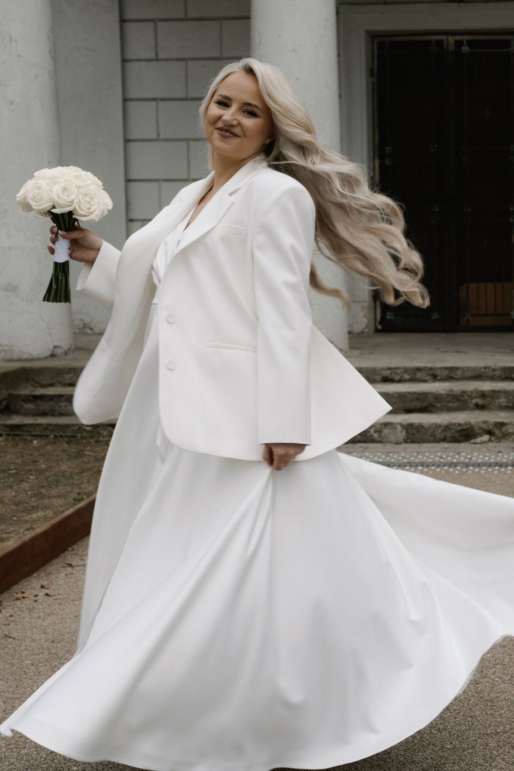 Bride in white blazer and skirt twirling with rose bouquet outside elegant building.