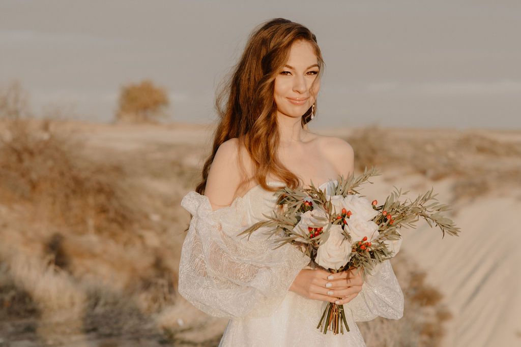 Bride in lace off-shoulder gown holding boho floral bouquet in golden desert landscape.