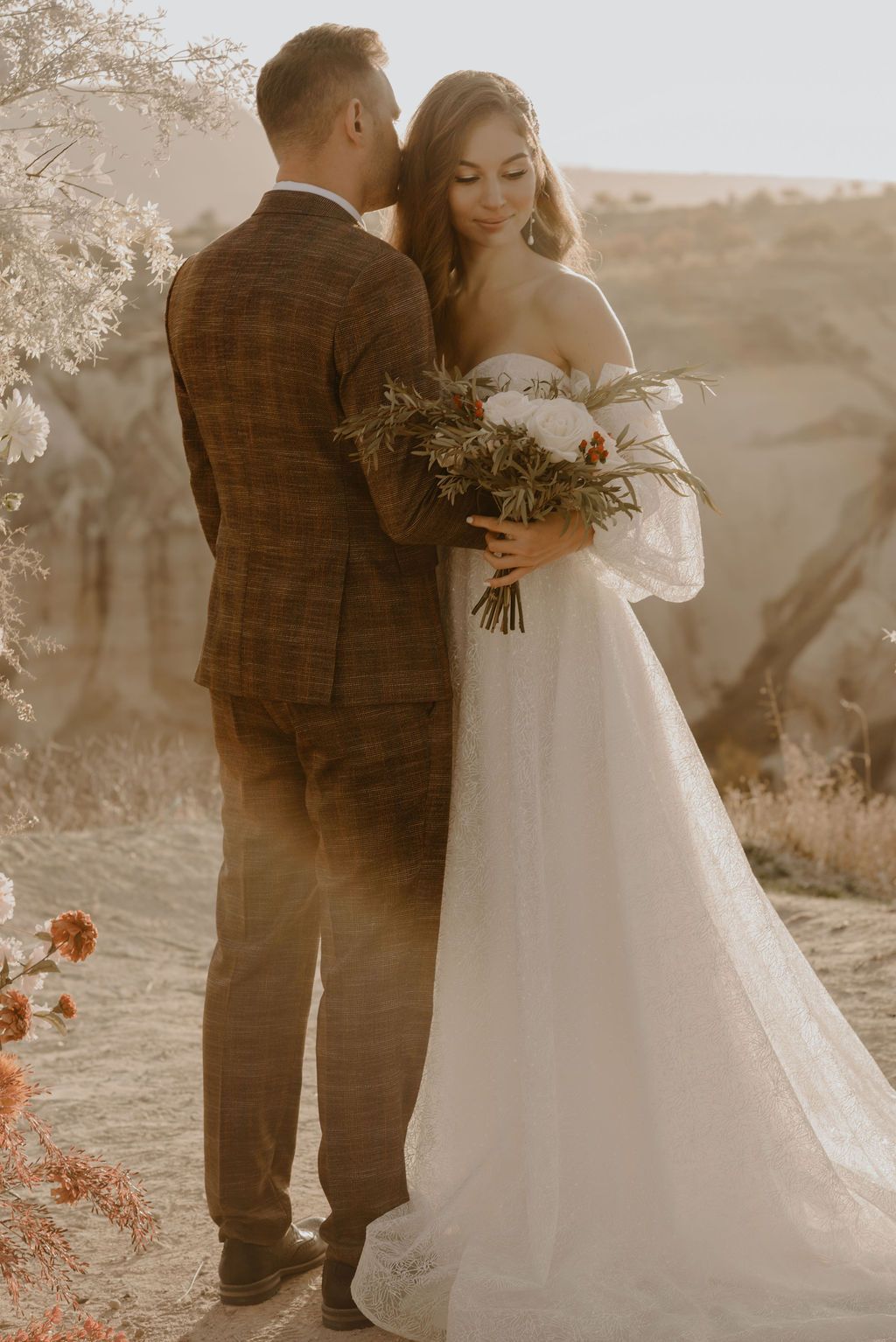 Bride in white off-shoulder gown holding bouquet stands with groom in plaid suit at outdoor wedding.
