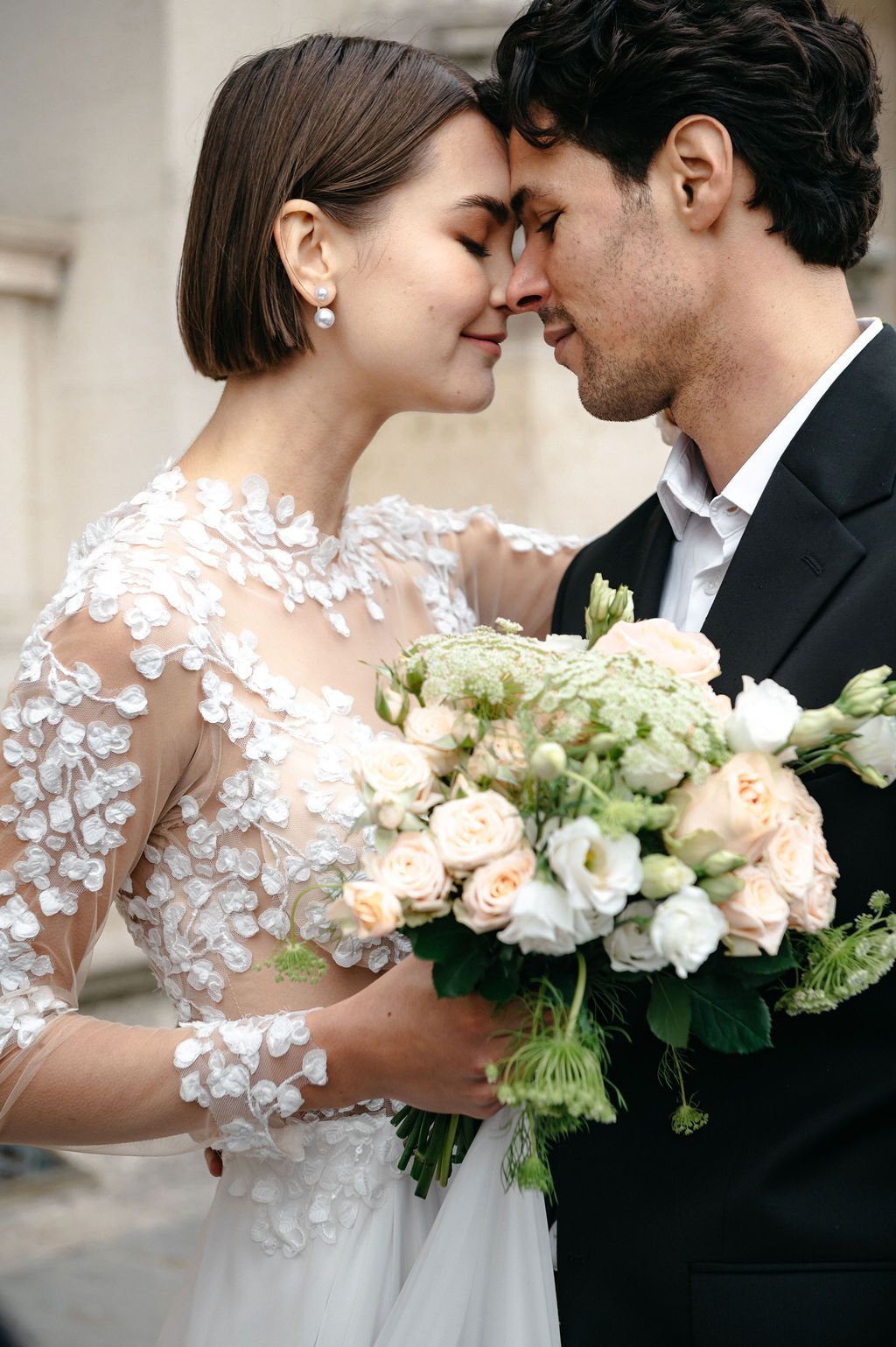 Bride and groom touching foreheads tenderly, bride holding elegant bouquet of white and blush roses in lace wedding dress.