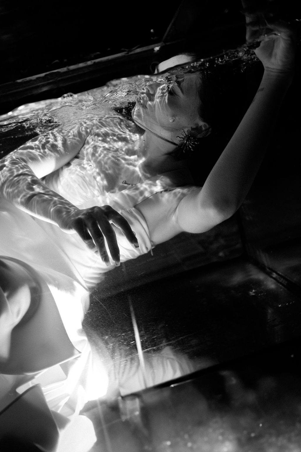 Black and white photo of a woman in white dress floating in water with dramatic lighting.