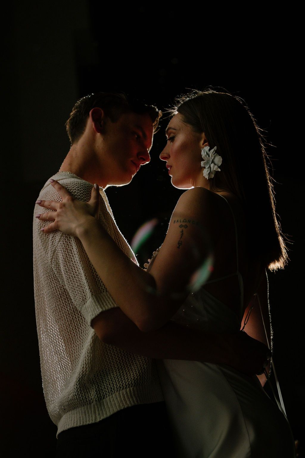 Romantic couple in dim lighting, woman with white flower in hair, embracing face-to-face in an intimate moment.