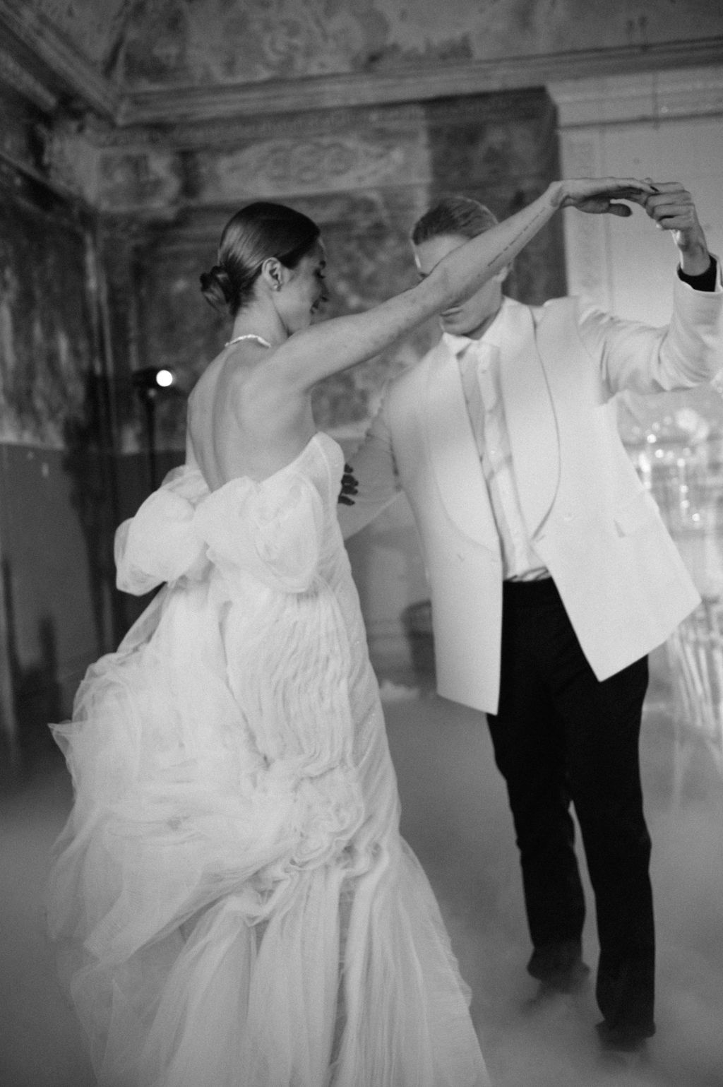 Black and white photo of bride and groom sharing first dance, bride in flowing gown, groom in white tuxedo jacket.