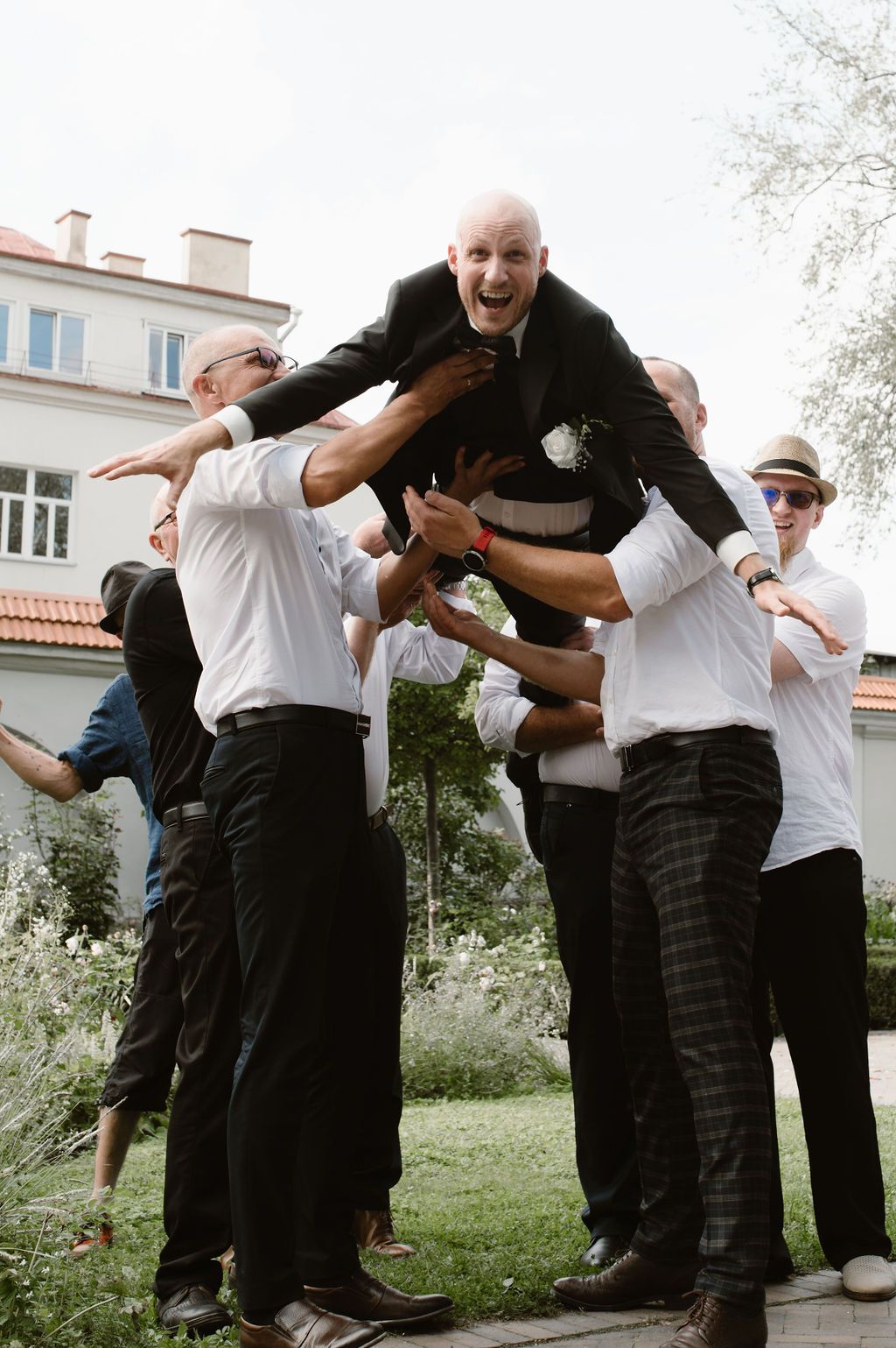 Groomsmen lifting a laughing bald groom in a black suit during an outdoor wedding celebration.
