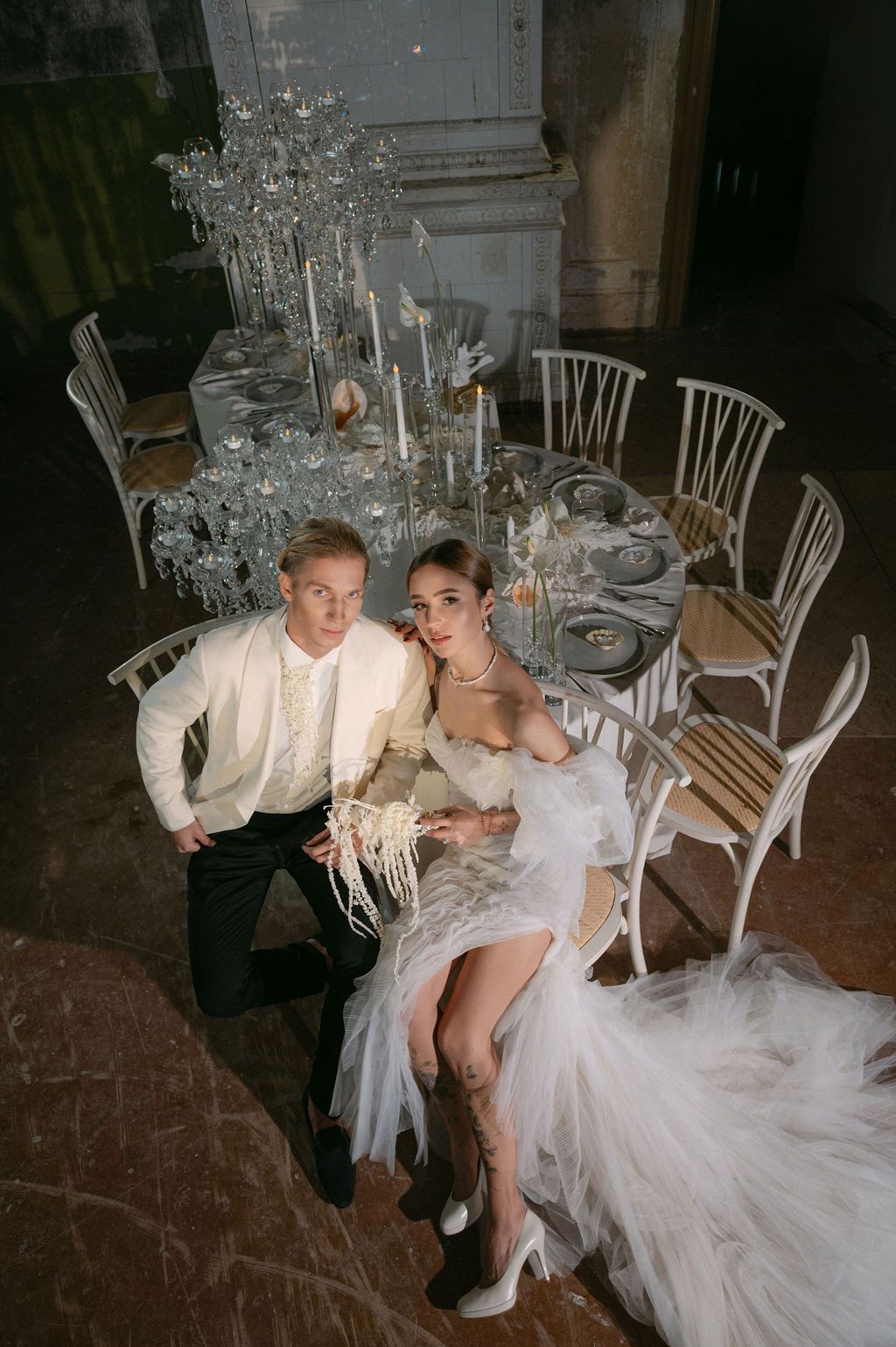 Elegant couple in wedding attire seated on floor beside decorated reception table with crystal candelabras in grand venue.