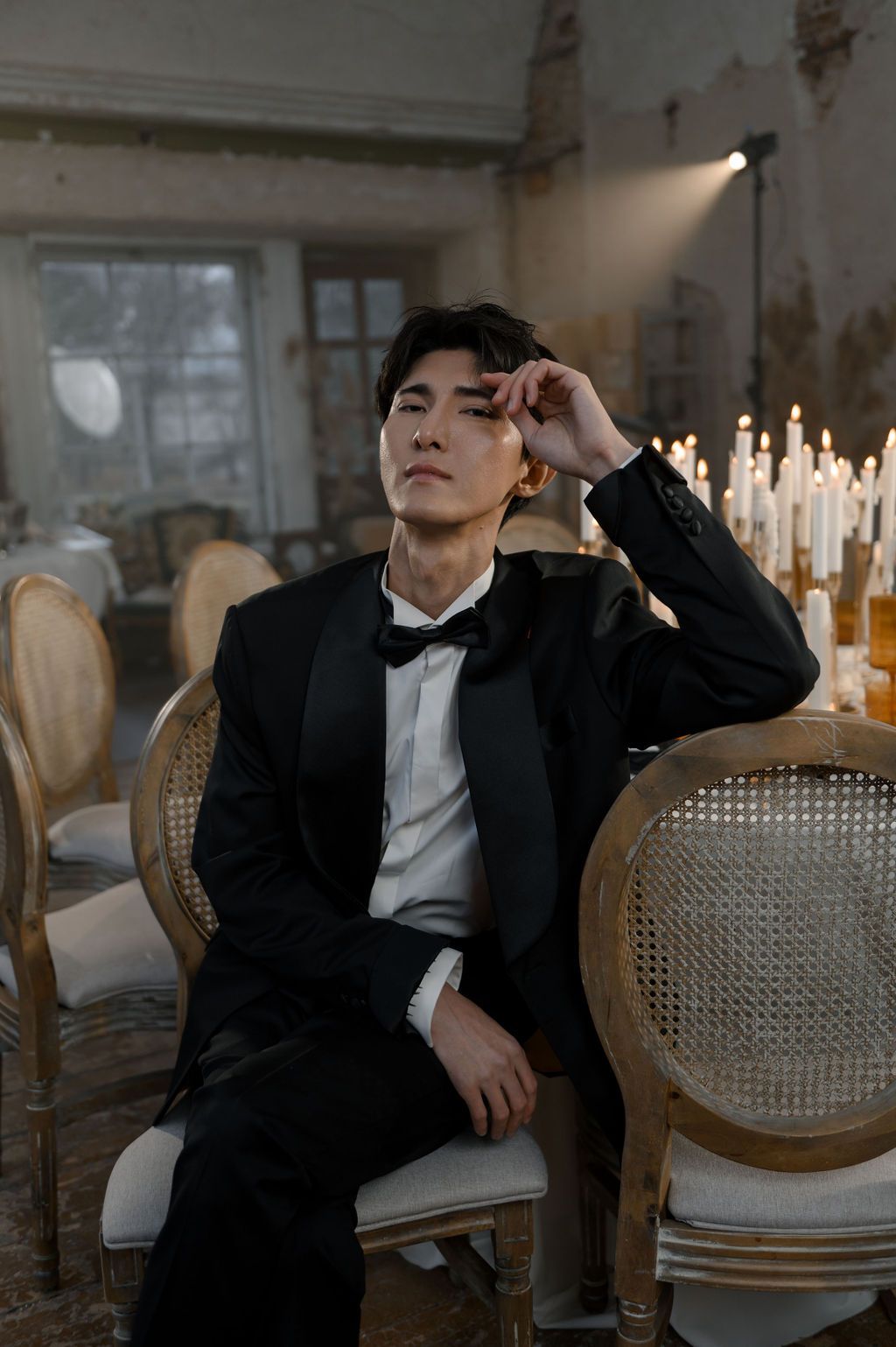 Man in black tuxedo sitting on ornate chair with candles in elegant room.