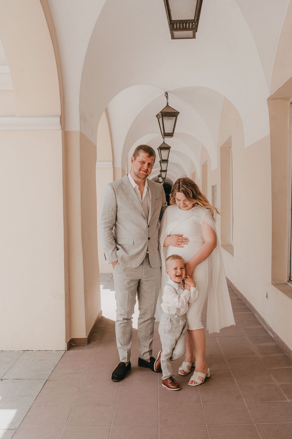 Pregnant woman in white dress poses with husband in gray suit and toddler in arched corridor.