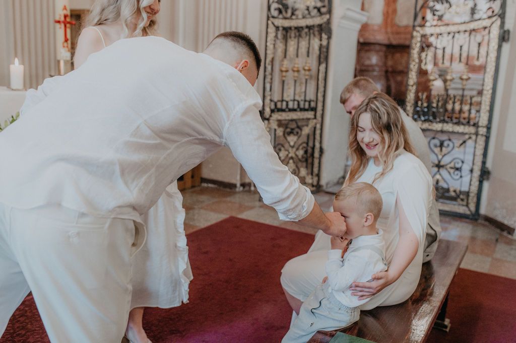 A priest performs a baptism ceremony on an infant held by a smiling mother inside a church.