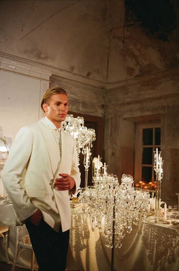 Groom in white tuxedo jacket stands beside crystal candelabras at elegantly decorated wedding reception table.