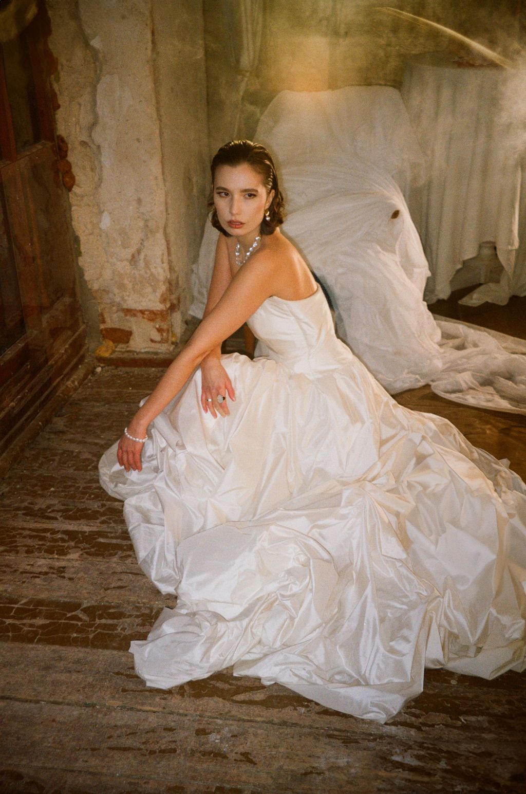 Bride in a strapless white ball gown sitting gracefully on a rustic wooden floor in an abandoned venue.
