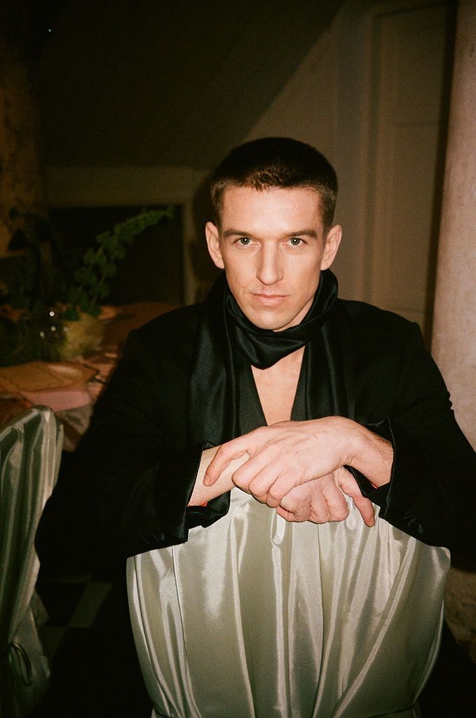 Young man in black jacket with scarf poses confidently leaning on silver fabric at an elegant evening event.