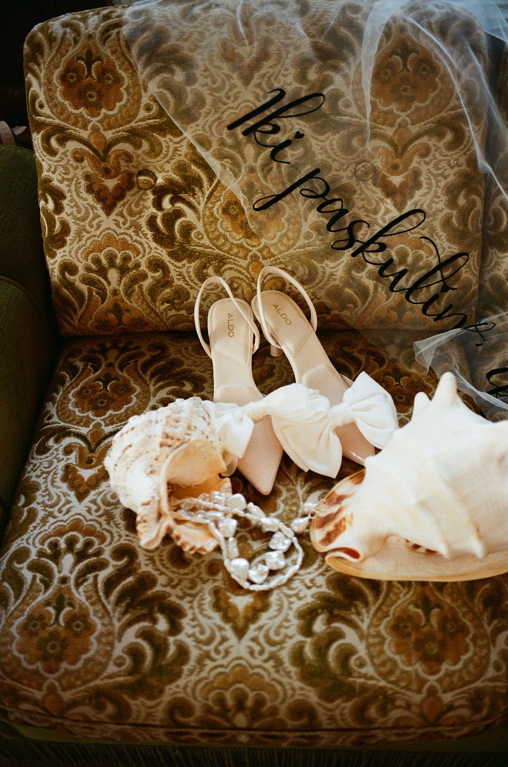 Bridal accessories on vintage floral chair featuring bow heels, pearl necklace, lace gloves, and veil for wedding day.