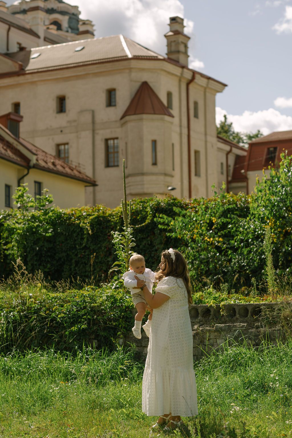 A mother in a white dress holds her baby outdoors near a historic European building surrounded by lush greenery.