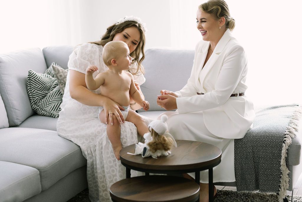 Two women in white outfits sit on a gray sofa with a baby and a small dog on a round coffee table.