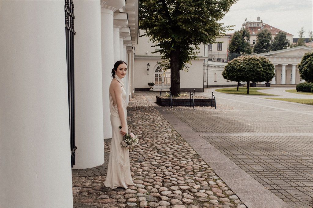 Bride in elegant white gown standing by white columns on cobblestone path at outdoor wedding venue.