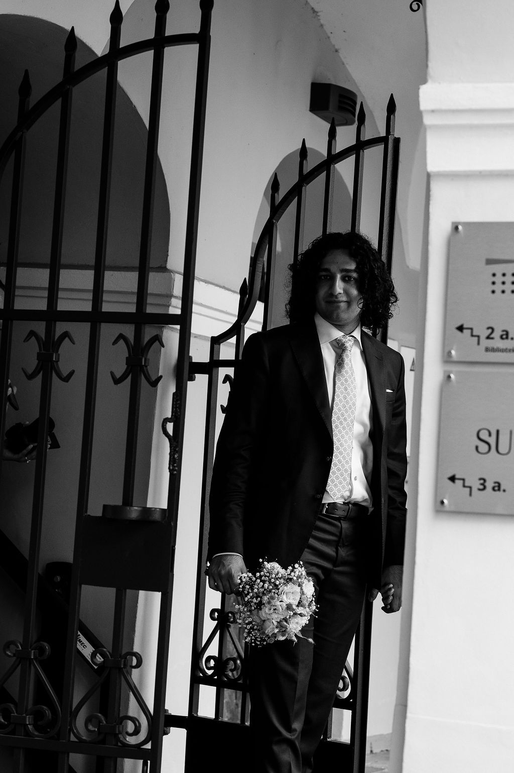 Groom in black suit holding white flower bouquet standing at ornate iron gate in black and white photo.