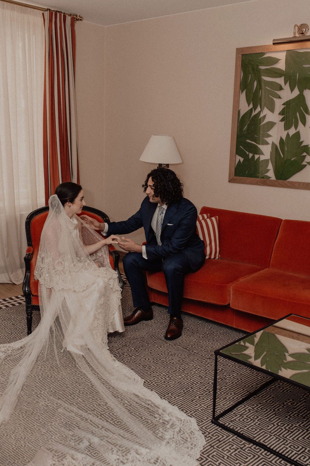 Bride in lace gown with cathedral veil holds hands with groom in navy suit on red velvet sofa in elegant hotel room.