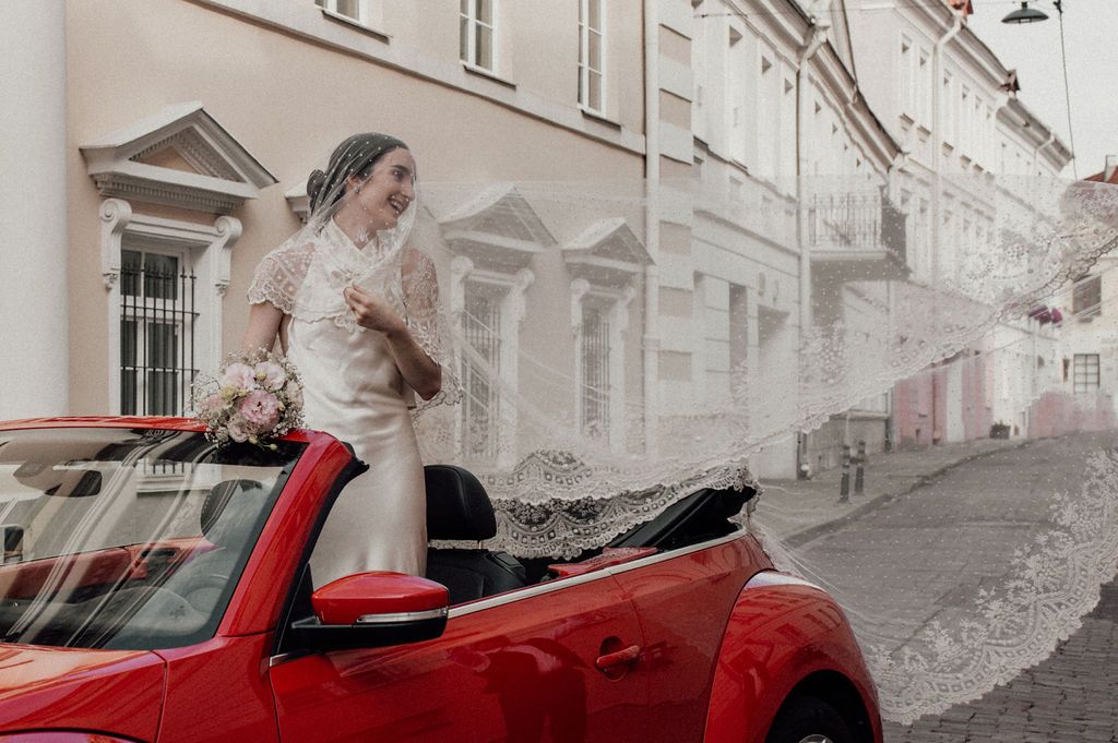 Bride in white wedding dress standing in red convertible car on a European cobblestone street holding a bouquet.